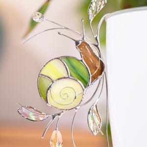 Close up of stained glass snail plant decor showing detailed green spiral shell and iridescent leaves. Crystal drops catch light, highlighting handcrafted glasswork.