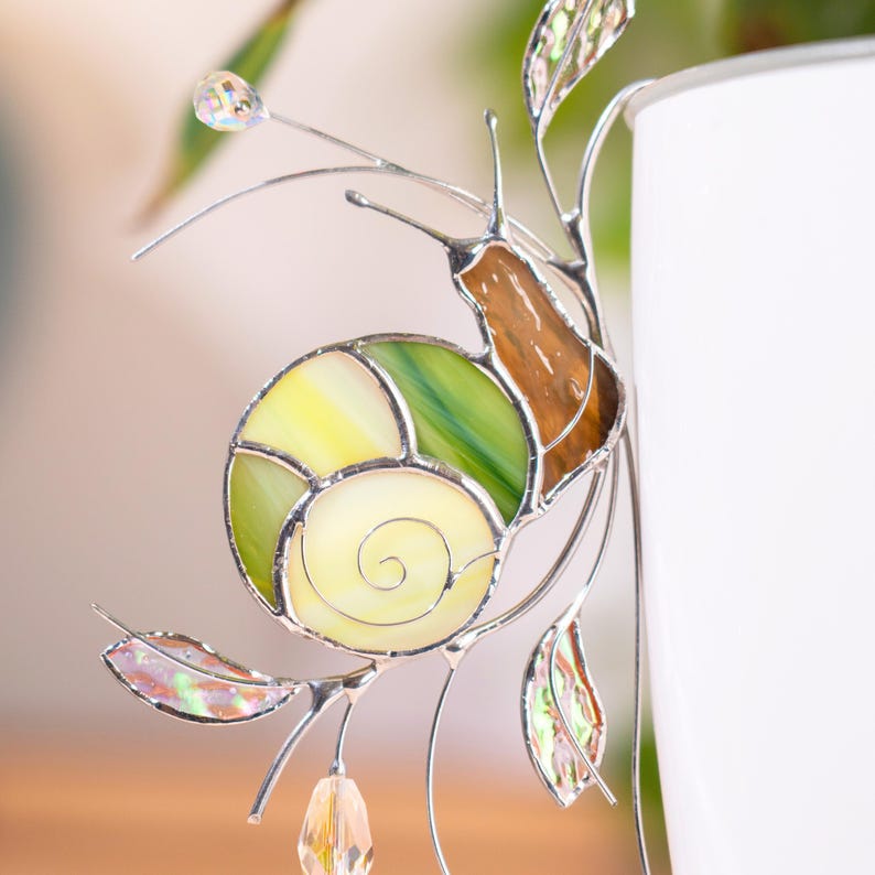 Close up of stained glass snail plant decor showing detailed green spiral shell and iridescent leaves. Crystal drops catch light, highlighting handcrafted glasswork.