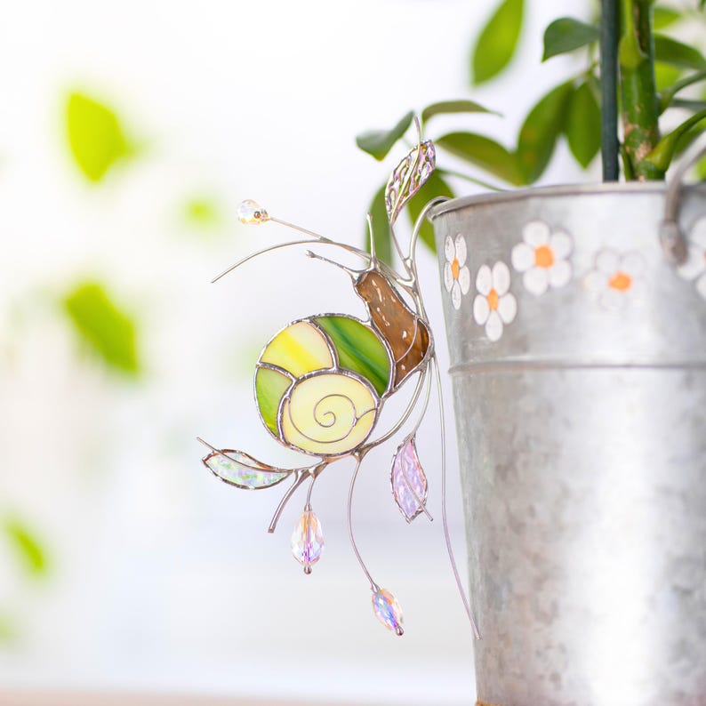 Stained glass snail suncatcher clipped to metal flower pot with daisy pattern, green shell and iridescent leaves, handmade glass plant decor with hanging crystal prisms.