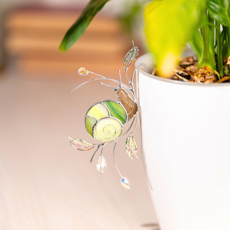 Stained glass snail plant pot decor hanging on white ceramic planter. Green and cream glass shell with delicate wire details and crystal beads adds whimsical botanical accent.