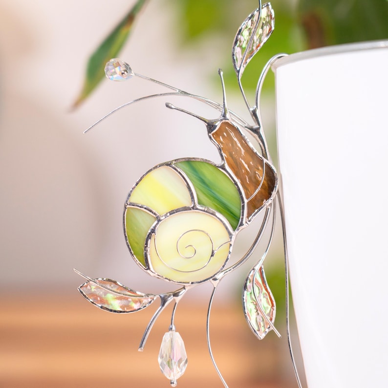 Stained glass snail plant decor clipped to white planter, handmade suncatcher with green spiral shell, amber body and crystal drops reflecting sunlight, decorative glass ornament for indoor plants.