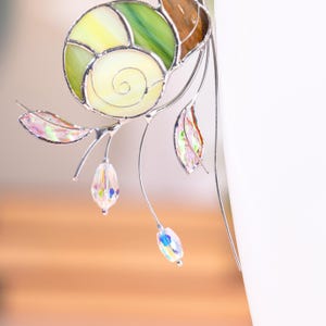 Macro close up of stained glass snail plant decor with spiral shell and dangling crystal prism. Sunlight enhances translucent glass textures and fine metalwork.