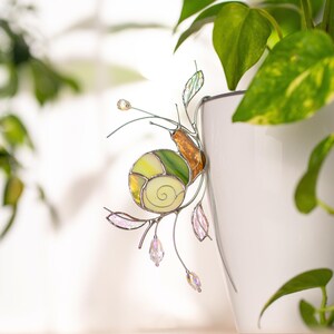 Stained glass snail plant decor on white pot with green houseplant, handmade suncatcher with translucent shell and crystal drops catching sunlight, botanical home decoration.