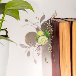 Stained glass snail plant decoration attached to book, green spiral shell suncatcher casting soft shadow, handcrafted glass ornament with crystal beads, cozy home decor accent.