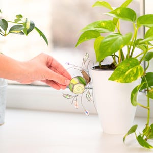 Stained glass snail decor being placed on plant pot, handmade suncatcher with green shell and crystal prisms, indoor plant accessory adding light reflections and artistic touch.