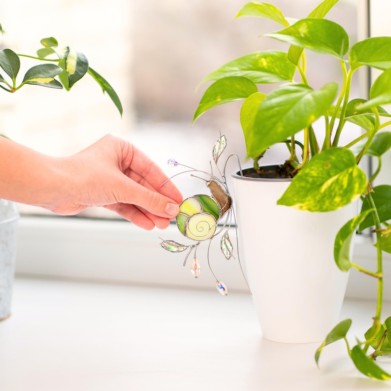 Stained glass snail decor being placed on plant pot, handmade suncatcher with green shell and crystal prisms, indoor plant accessory adding light reflections and artistic touch.