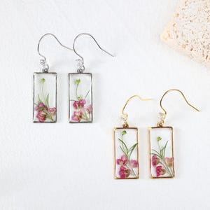 May include: Earrings with rectangular resin pendants, each showcasing pink flowers and green stems. Two pairs are visible: one with silver-tone hooks and frames, the other with gold-tone hooks and frames. The earrings are on a white background.