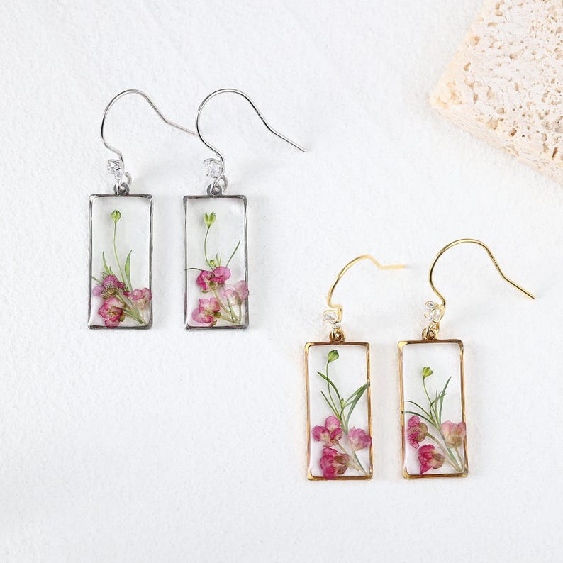May include: Earrings with rectangular resin pendants, each showcasing pink flowers and green stems. Two pairs are visible: one with silver-tone hooks and frames, the other with gold-tone hooks and frames. The earrings are on a white background.