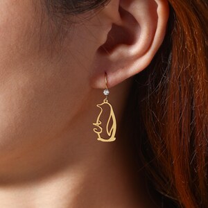 May include: Gold-colored penguin earrings with a small, clear gemstone above the penguin. The earrings are hanging from a person's ear. The penguin design is a stylized outline.
