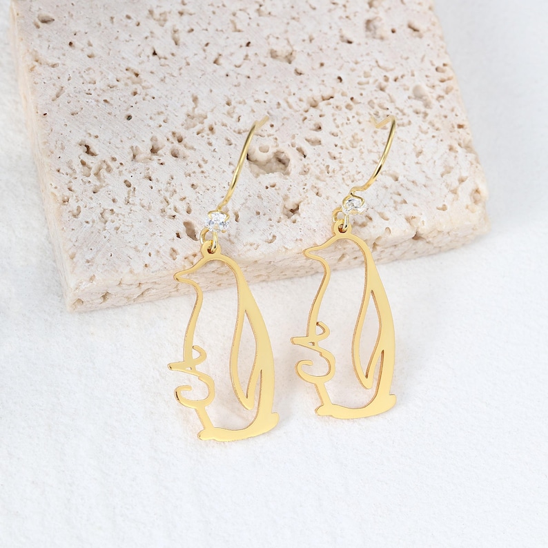 May include: Gold-colored penguin-shaped dangle earrings. Each earring features a cutout design of a penguin with a small, clear gemstone at the top. The earrings are displayed on a textured, light-colored surface.