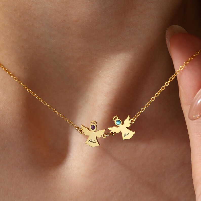 May include: A gold-colored necklace featuring two angel pendants. Each angel has a gemstone halo and a name engraved below. The necklace is worn around the neck, with the chain visible.