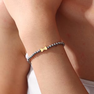May include: A beaded bracelet with small, silver-coloured beads. A gold-coloured charm in the shape of a dog bone is centered on the bracelet, with the name "Ace" engraved on it. The bracelet is worn on a wrist.