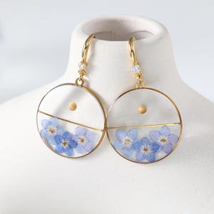 May include: Gold-tone hoop earrings with clear resin circles. Each earring features a horizontal gold bar, blue forget-me-not flowers, and a small wooden bead. The earrings have a small, clear gem at the top.