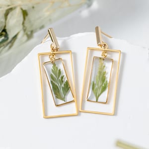 May include: Gold-toned rectangular earrings. Each earring features a clear, rectangular resin pendant with a green fern leaf encased inside. The pendants are suspended within a larger, open gold rectangle, creating a layered effect.