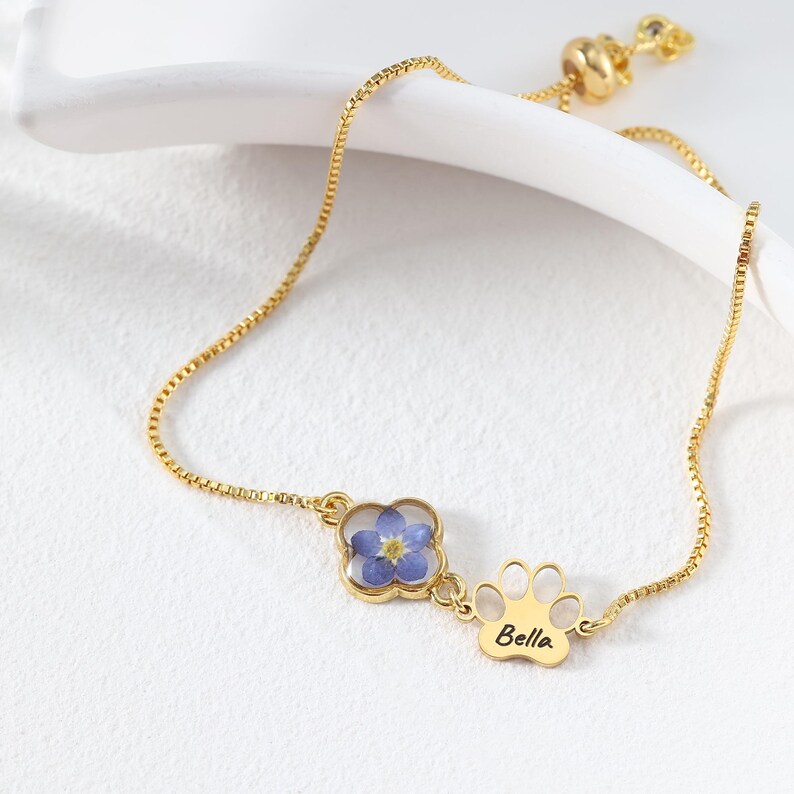 Puede incluir: Una pulsera dorada con una delicada cadena. Cuenta con un dije de tr&eacute;bol de cuatro hojas con una flor azul y un dije en forma de pata con el nombre "Bella" grabado. La pulsera tiene un cierre deslizante ajustable.
