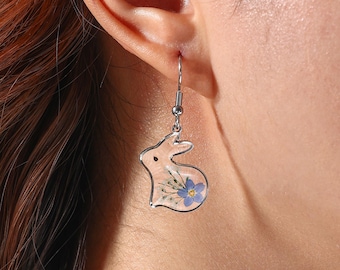 May include: A silver-toned earring featuring a bunny-shaped pendant. The pendant is filled with dried flowers, including a blue flower. The earring is hanging from a person's ear, showcasing the delicate design and floral elements.