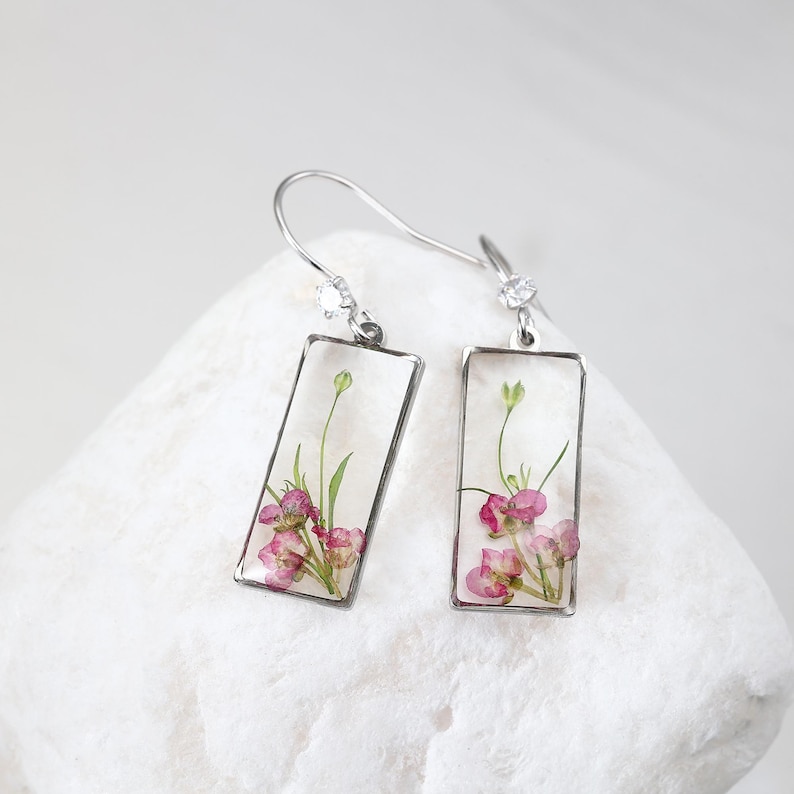May include: Silver-tone dangle earrings with rectangular clear resin pendants. Each pendant contains small pink flowers and green stems. The earrings have a hook closure with a small clear gem.