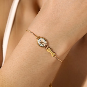 Könnte beinhalten: Ein goldfarbenes Armband mit einem ovalen Anhänger, der ein Hundeporträt zeigt. Ein knochenförmiger Anhänger mit dem Wort "Caesar" ist angebracht. Das Armband wird am Handgelenk getragen.