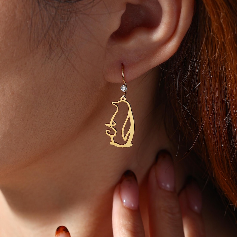 May include: Gold-colored penguin earrings with a small, clear gemstone at the top. The earrings are dangling from a person's ear. The penguin design is a simple outline.