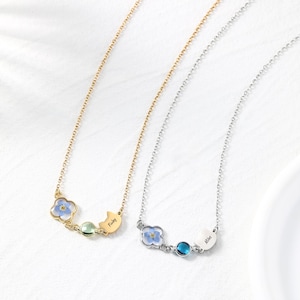 May include: Two necklaces with floral and gemstone pendants. One is gold-toned, with a blue flower, a green gem, and a charm with "Finley." The other is silver-toned, with a blue flower, a blue gem, and a charm with "Miles."