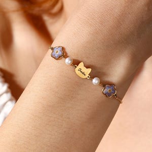 Pode incluir: Uma pulseira dourada com um pingente em forma de gato com a inscrição "Shadow", intercalada com pequenas pérolas e pingentes de flores roxas claras. A pulseira é delicada e elegante.
