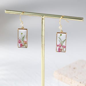 May include: Gold-tone dangle earrings with rectangular clear resin pendants. Each pendant features pink flowers and green stems. A small clear crystal bead is attached to the top of each earring.