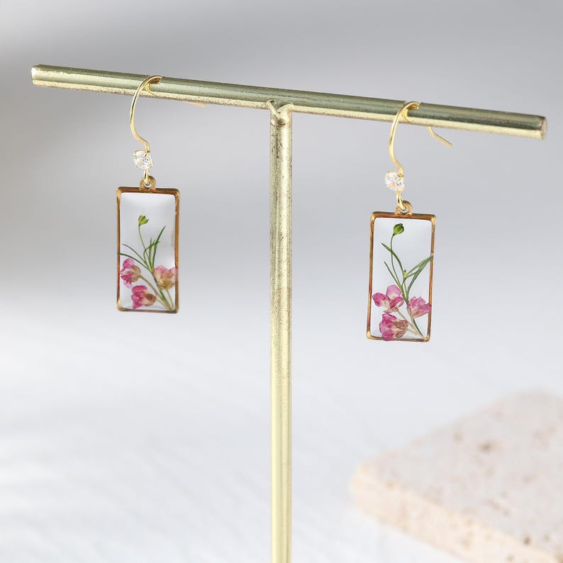 May include: Gold-tone dangle earrings with rectangular clear resin pendants. Each pendant features pink flowers and green stems. A small clear crystal bead is attached to the top of each earring.