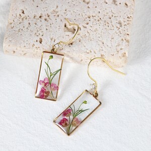 May include: A pair of gold-colored rectangular earrings with a clear resin center, showcasing dried pink flowers and green stems. Each earring has a small, clear gem and a curved hook.