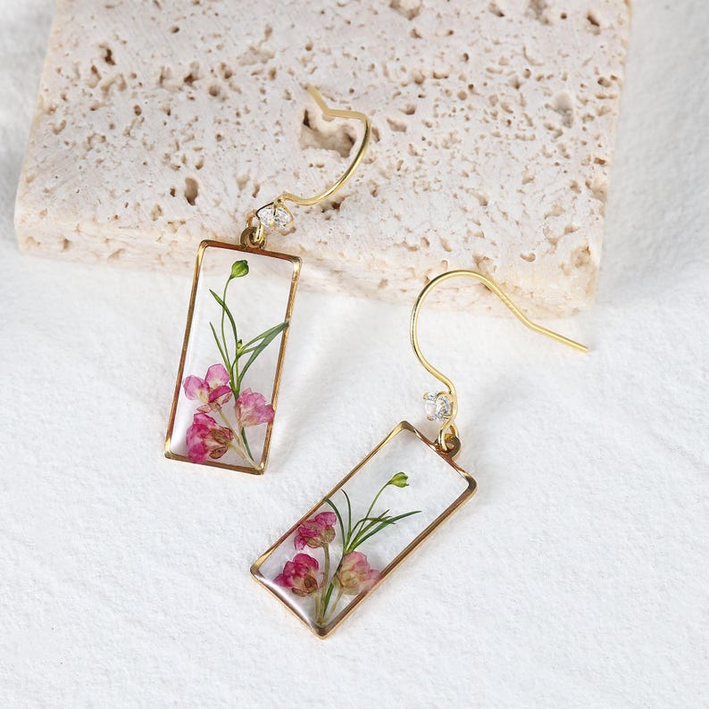 May include: A pair of gold-colored rectangular earrings with a clear resin center, showcasing dried pink flowers and green stems. Each earring has a small, clear gem and a curved hook.
