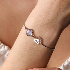 Puede incluir: Una pulsera plateada con un dije de flor y un dije de huella de pata con el nombre "Mia" grabado. El dije de flor tiene un dise&ntilde;o floral azul y morado. La pulsera se lleva en la mu&ntilde;eca.