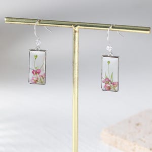 May include: Silver-tone dangle earrings with rectangular clear resin pendants. Each pendant features a delicate arrangement of pink and green dried flowers. The earrings are displayed on a gold-colored jewelry stand.