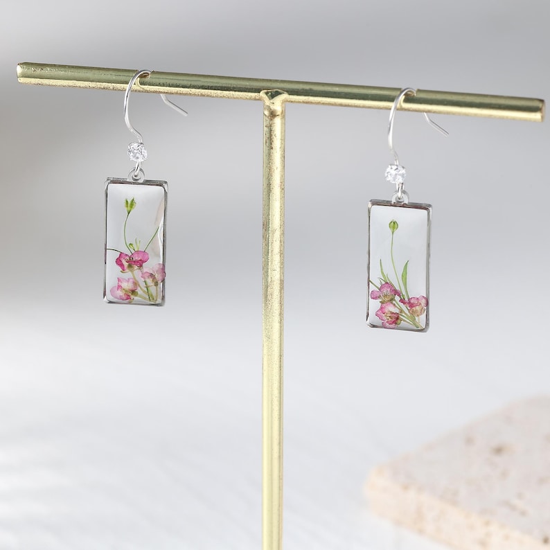 May include: Silver-tone dangle earrings with rectangular clear resin pendants. Each pendant features a delicate arrangement of pink and green dried flowers. The earrings are displayed on a gold-colored jewelry stand.