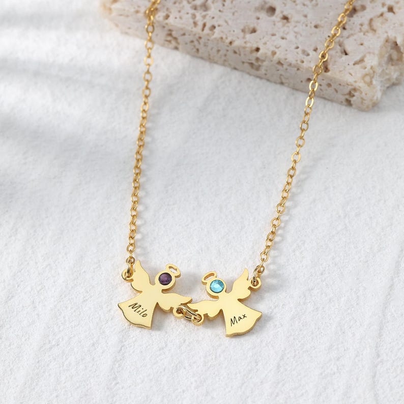 May include: Gold-colored necklace featuring two angel pendants. Each angel has a halo with a gemstone and a name engraved on the body. The necklace is on a white surface.