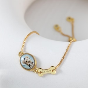 Op de afbeelding: Goudkleurige verstelbare armband met een ovale hanger met een hondenportret en een botvormige bedel met de inscriptie "Caesar". De armband heeft een delicate ketting en een schuifsluiting.