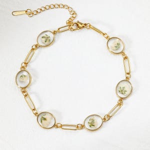 May include: Gold-toned bracelet featuring oval-shaped clear resin settings, each containing a small, dried white flower. The bracelet has a delicate chain design with a lobster clasp closure. The overall aesthetic is elegant and nature-inspired.