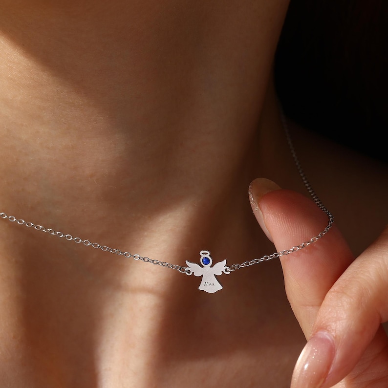 May include: A silver angel pendant necklace with a delicate chain. The angel has a blue gem and the name "Max" engraved on it. The necklace is being worn around a person's neck.