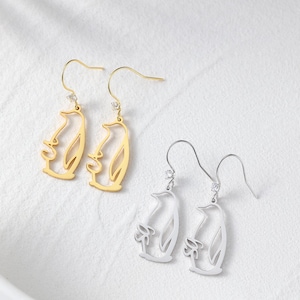 May include: Gold and silver-colored penguin-shaped dangle earrings. The earrings feature a cutout design of a penguin with a small gemstone at the top. The gold earrings have a polished finish, while the silver earrings have a matte finish.