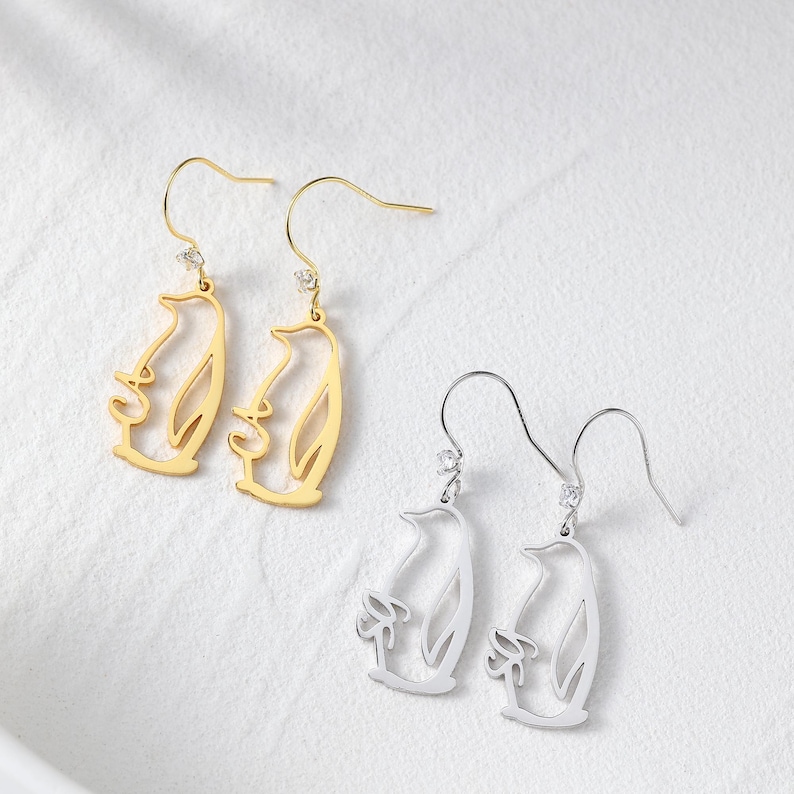 May include: Gold and silver-colored penguin-shaped dangle earrings. The earrings feature a cutout design of a penguin with a small gemstone at the top. The gold earrings have a polished finish, while the silver earrings have a matte finish.