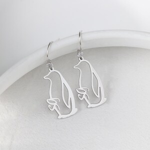 May include: A pair of silver-colored dangle earrings. Each earring features a cutout design of a penguin holding a fish. The earrings have a small, clear gem at the top and a curved hook for the ear.