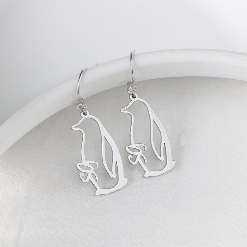 May include: A pair of silver-colored dangle earrings. Each earring features a cutout design of a penguin holding a fish. The earrings have a small, clear gem at the top and a curved hook for the ear.
