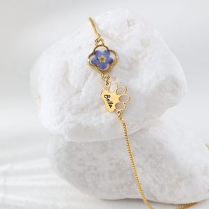 Puede incluir: Un collar dorado con un colgante de tr&eacute;bol de cuatro hojas con una flor azul y un dije en forma de pata con el nombre "Bella". El collar se muestra sobre una superficie blanca texturizada.