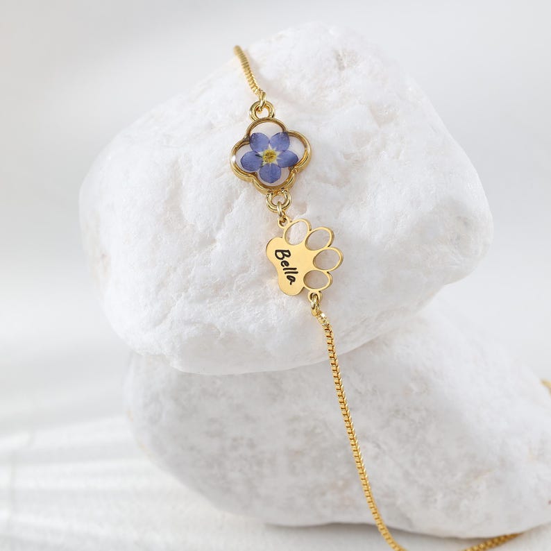 Puede incluir: Un collar dorado con un colgante de tr&eacute;bol de cuatro hojas con una flor azul y un dije en forma de pata con el nombre "Bella". El collar se muestra sobre una superficie blanca texturizada.