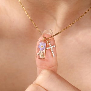 May include: A gold-colored necklace featuring two pendants. One pendant is a rectangular clear resin piece with a purple flower and white flowers inside. The other pendant is a small cross adorned with clear crystals.