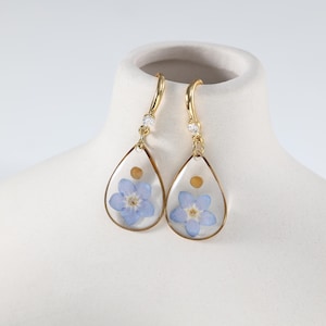 May include: Gold-colored dangle earrings with a teardrop shape. Each earring features a light blue flower encased in clear resin, with a small gold-colored accent and a sparkling clear gem at the top. The earrings are displayed on a white surface.
