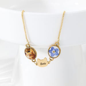 May include: A gold-toned chain necklace featuring three circular and cat-shaped pendants. One pendant displays a photograph of an orange tabby cat, another contains a blue forget-me-not flower, and the centre cat-shaped charm is engraved with the name Boots.