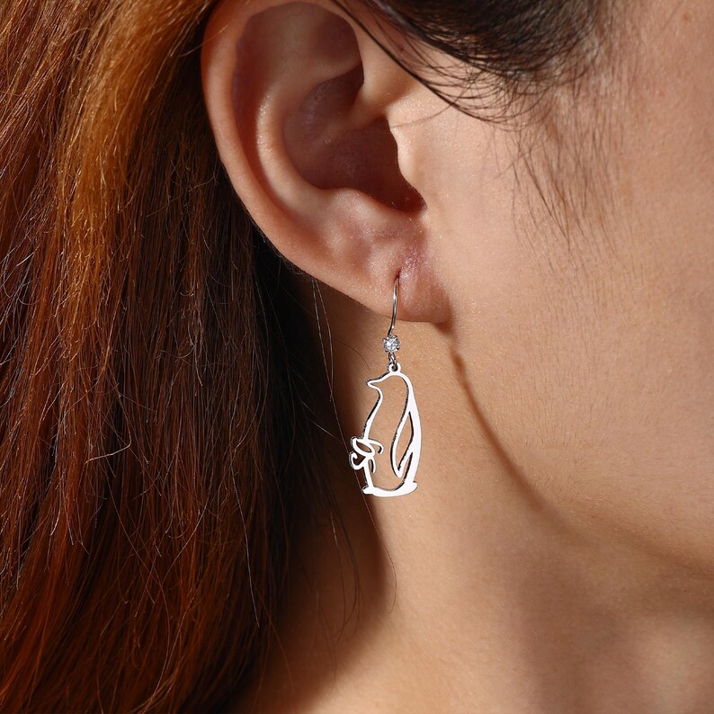 May include: Silver penguin dangle earrings. The earrings feature a cut-out penguin design with a small, sparkling accent above the penguin. The earrings are suspended from a simple hook.