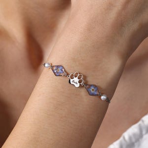 May include: Silver bracelet with a paw print charm inscribed with "Bella" and two diamond-shaped charms containing blue flowers. The bracelet is accented with small pearls.