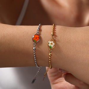 May include: Two bracelets with floral designs. One bracelet is silver-toned with a chain of small beads and a circular pendant with an orange flower. The other is gold-toned with a chain of small beads and a circular pendant with a white flower.
