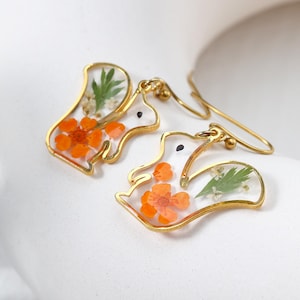 Op de afbeelding: Eikelvormige oorbellen met goudkleurige frames. De met heldere hars gevulde lichamen bevatten oranje bloemen, groene bladeren en kleine witte bloemen. De oorbellen hebben goudkleurige oorhaken en kleine gouden kralen.