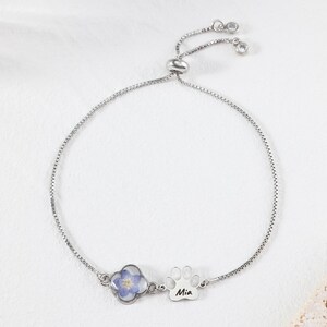 Puede incluir: Pulsera ajustable plateada. Presenta un tr&eacute;bol de cuatro hojas con una flor azul y un dije de huella de pata con el nombre "Mia". La pulsera tiene una cadena delicada y un cierre deslizante.